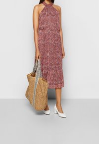 Frau in einem ärmellosen, pinkfarbenen, blumigen Halterneck-Kleid, mit weißen Blockabsätzen und einer großen, geflochtenen Tote-Tasche mit gemusterten Trägern.