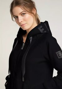 Schwarzer Hoodie mit Reißverschluss, strukturierten Kordeln und Logo-Details an Kapuze und Ärmeln. Glattes Material mit taillierter Passform.