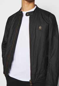 Czarna kurtka bomber z zamkiem z przodu, zaokrąglonym kołnierzem i zatrzaskami na guziki. Posiada subtelne detale z logo i gładką teksturę.