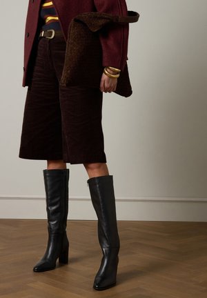 Zwarte leren laarzen tot over de knie met een vierkante neus en een stevige hak. Draagt ze bij een bruine corduroy korte broek en een bruin gestructureerde tas.