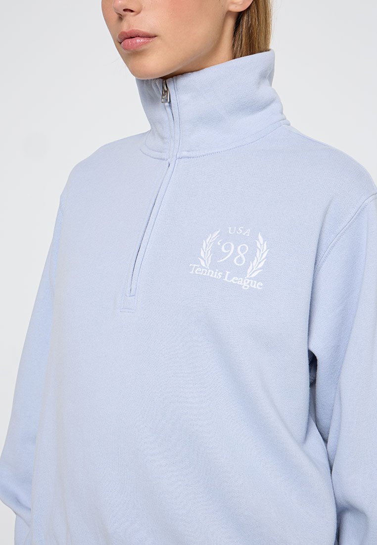 Helle blaue Zip-Up Sweatshirt aus weichem Material, mit einem gestickten Logo "USA 98 Tennis League" in Weiß auf der Vorderseite.