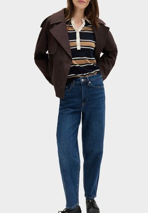 Femme portant une veste courte marron foncé sur un polo rayé, un jean bleu taille haute et des chaussures noires, debout les mains derrière le dos.