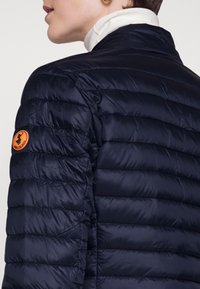 Granatowa kurtka puchowa z poziomym pikowanym wzorem, z pomarańczową naszywką z logo na lewym ramieniu i wysokim kołnierzem. Gładka faktura.