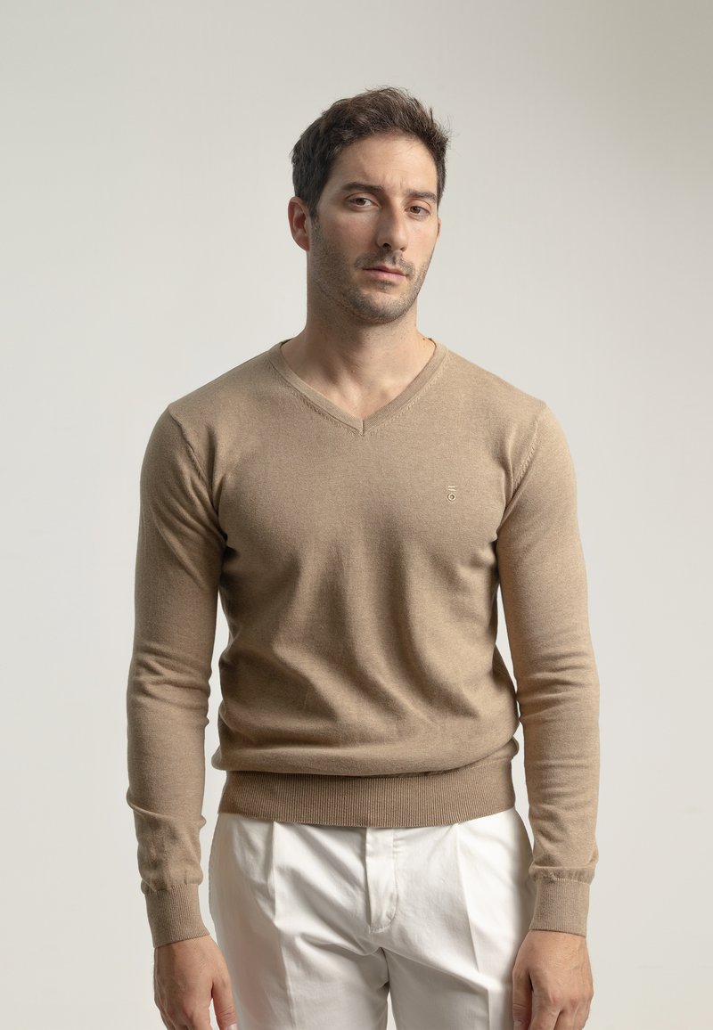 Suéter de punto beige con escote en V, mangas largas y puños acanalados; presenta un pequeño logo en el lado izquierdo del pecho. Combinado con pantalones blancos.