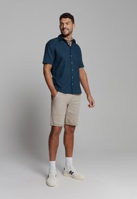 Korte mouwen, donkerblauwe overhemden met knopen, beige shorts, witte sneakers met marineblauwe accenten, staande pose, handen in de zakken, casual stijl.