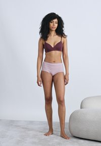 Καφέ σουτιέν με λεπτή ταινία και σχεδιασμό σε σχήμα V, σε συνδυασμό με ροζ ανοιχτό, ψηλόμεσο εσώρουχο. Το μοντέλο στέκεται σε υφή γκρι δαπέδου.