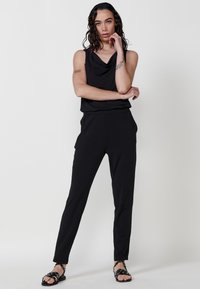 Črno kombinezon s drapiranim ovratnikom, slim fit, stranskima žepoma in dolžino do gležnjev. Oblečen z ravnimi črnimi sandali. Gladek, lahek material.