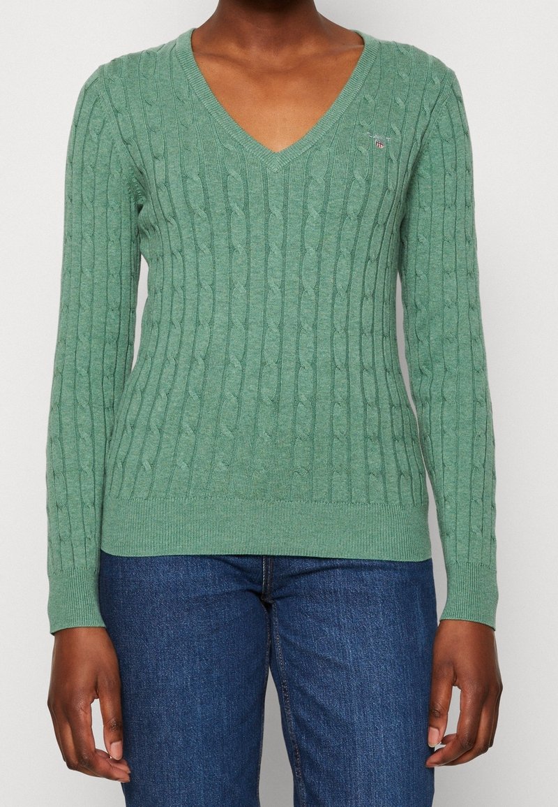 Personne portant un pull vert à col en V en tricot torsadé et un jean bleu, debout devant un fond clair uni.