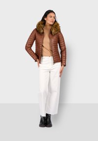 Bruin gewatteerd pufferjack met een faux fur capuchon, gecombineerd met een beige turtleneck en witte wijde broek. Zwarte enkellaarzen maken de look compleet.