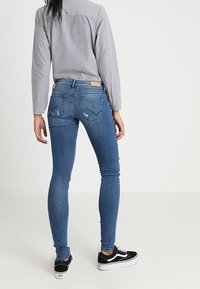 Jeans ajustados en denim azul medio, con una cintura alta, sutiles bolsillos traseros y una textura suave, combinados con zapatillas negras.