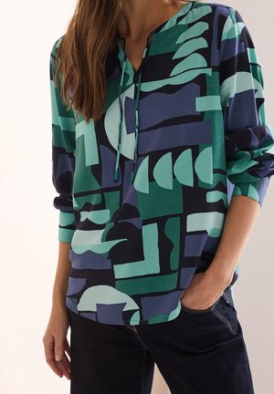 Femme portant un chemisier à manches longues avec des motifs géométriques abstraits bleus, verts et noirs et un pantalon foncé, main dans la poche.