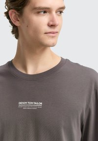 Pelēka garām piedurknēm T-krekls, izgatavots no mīkstas kokvilnas, ar apaļu kakla izgriezumu un balti drukātu logotipu uz krūtīm. Gluda tekstūra ar vienkāršu dizainu.