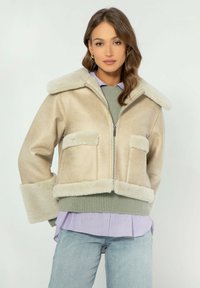 Giacca in shearling beige con ampio colletto e zip, dotata di due tasche frontali e orlo verde a coste. Indossata sopra una camicia lilla chiaro.