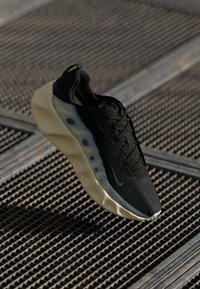 Pantofii atletici negri, cu un superior texturat, guler căptușit și o talpă translucidă de crem. Au un design aerodinamic și un branding minim.