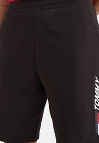Shorts neri in cotone con vita elasticizzata, tasche laterali e logo bianco "TOMMY" con accenti rossi e blu lungo il lato.