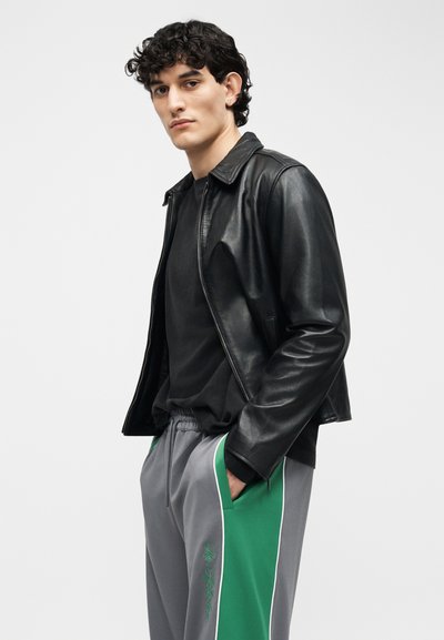 Νέος άνδρας με σγουρά μαλλιά, φορώντας μαύρο δερμάτινο μπουφάν, μαύρο πουκάμισο και γκρι-πράσινη φόρμα με τα χέρια στις τσέπες.