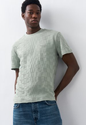 Jeune homme portant une chemise à manches courtes texturée vert clair et un jean bleu, debout contre un mur blanc, les mains derrière le dos.