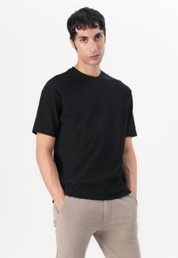 Tricou negru cu mânecă scurtă, dintr-un material texturat, cu decolteu rotund și croi lejer. Asortat cu pantaloni bej, pe corp.