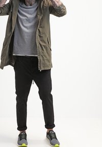 Man i olivfärgad jacka, grå t-shirt, svarta uppvikta byxor, grå och neongröna sneakers, med tatuerade underarmar höjda nära huvudet.