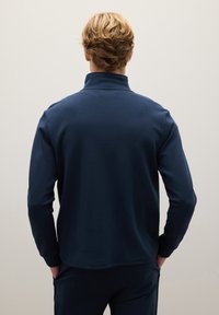 Sudadera azul marino con cuello alto, textura suave y mangas largas. El diseño se sitúa a la altura de las caderas y ofrece un ajuste entallado sin logotipos.