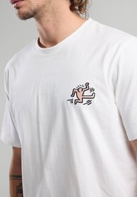 White cotton t-shirt featuring a pink and black embroidered design of a running figure on the left chest area. Round neckline, short sleeves.