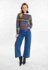 Mehrfarbiger gestrickter Pullover mit horizontalen Streifen in Lila, Grün und Braun; kombiniert mit hochgeschnittenen blauen Jeans-Hosen mit weitem Bein und schwarzen Stiefeln.