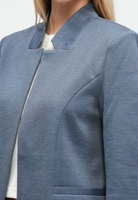 Modrý blejzer vyrobený z textúrovanej látky s vysokým golierom a priliehavým strihom. Má jednoradové zapínanie vpredu a bočné vrecká.