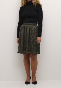 Haut à manches longues noir associé à une jupe plissée longueur genoux avec un motif floral vert foncé et taille élastique. Chaussures noires à talons pointus.
