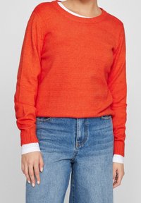 Pull tricoté orange vif avec un col rond et des poignets côtelés, associé à un jean taille haute bleu clair, avec un détail blanc sur la manche.