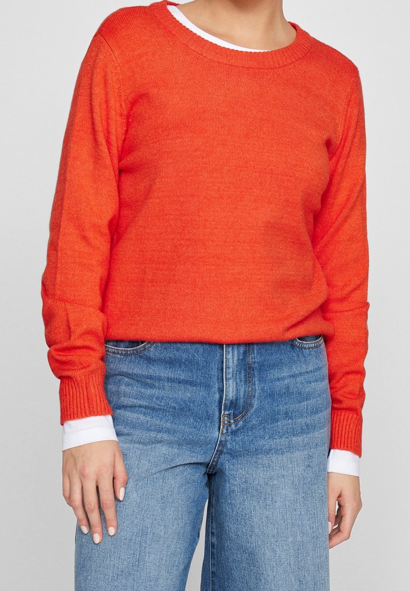 Pull tricoté orange vif avec un col rond et des poignets côtelés, associé à un jean taille haute bleu clair, avec un détail blanc sur la manche.