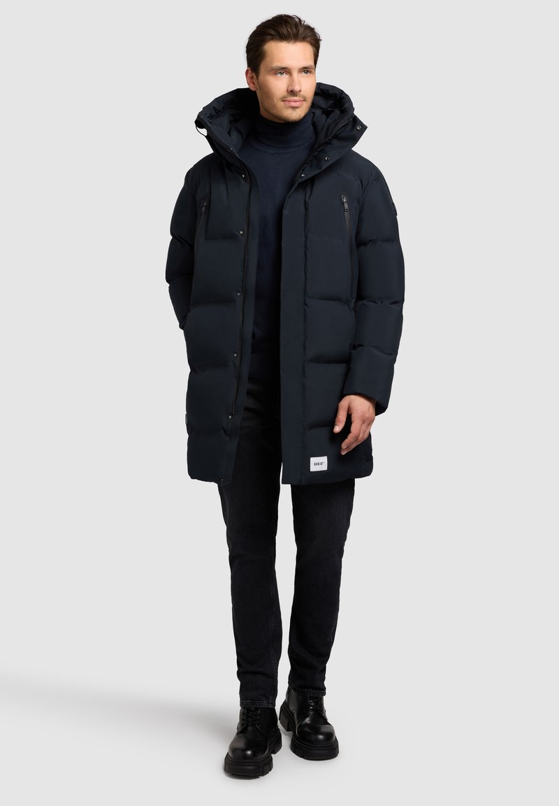 Marineblå puffjakke med aftagelig hætte, lynlåslommer, tekstureret stof og en afslappet pasform, parret med mørke jeans og sorte sko.