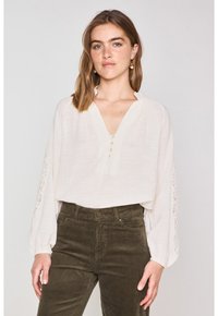 Blusa bianca con scollo a V e pattina dei bottoni, caratterizzata da dettagli in pizzo sulle maniche. Abbinata a pantaloni in velluto a coste color olivo.