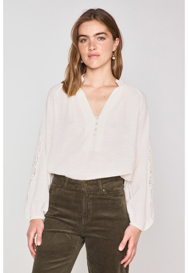 Blusa bianca con scollo a V e pattina dei bottoni, caratterizzata da dettagli in pizzo sulle maniche. Abbinata a pantaloni in velluto a coste color olivo.