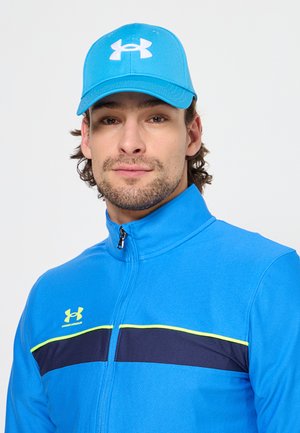 Man wearing a blue Under Armour cap and matching zip-up jacket with black and neon yellow stripes.