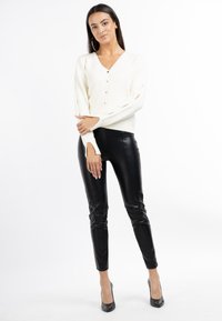 Maglione a maglia crema con scollo a V e chiusura con bottoni, abbinato a pantaloni in ecopelle neri. Modella in tacchi alti neri si trova davanti a uno sfondo bianco.
