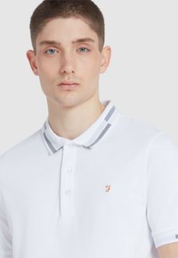 Polo bianco con colletto a righe, logo ricamato arancione e tessuto testurizzato. Presenta tre bottoni sul colletto.