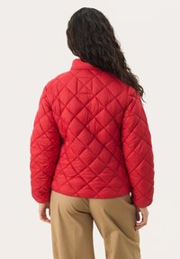 Red quilted jacket with a high collar and diamond pattern, featuring puffy insulation and a relaxed fit. Paired with beige trousers.