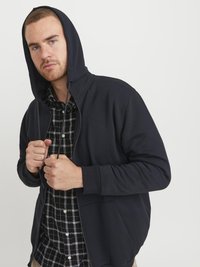 Mornarska zip-up dukserica s podstavljenom kapuljačom, prednjim džepovima i rebrastim manžetama, složena preko crno-bijele karirane košulje.