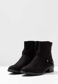 Botas de tobillo de ante negro con cierre de cremallera, detalle de correa envuelta y acentos metálicos decorativos, con un tacón bajo apilado.