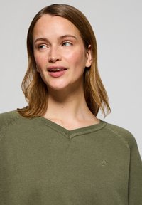 Olivgrön v-ringad tröja med ribbad textur, som har en synlig logga på bröstet och en avslappnad passform, framträdande med insydda ärmar.