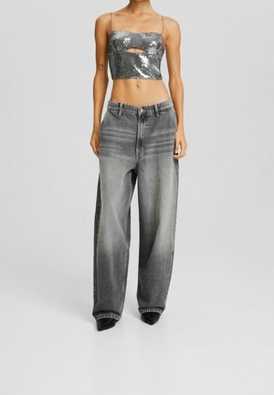 Mujer con un top corto plateado de lentejuelas con tirantes finos y vaqueros gris desgastados de pierna ancha, parada frente a un fondo liso.