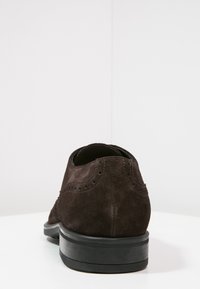 Chaussure en daim marron avec détails perforés, talon arrondi et semelle en caoutchouc noire. Vue arrière mettant en valeur la texture lisse et les accents de couture.