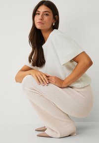 Hvid kortærmet sweater, strukturerede lyserøde strikkede bukser og beige spidse hæle, poserende i en squat-holdning mod en neutral baggrund.