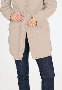 Veste imperméable beige avec fermetures à bouton et à zip, poignets réglables et deux poches avant, portée sur un jean bleu foncé.