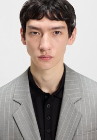 Young man with short black hair wearing a light gray pinstripe blazer over a black buttoned shirt, facing forward with a neutral expression.