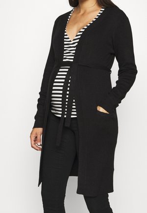 Femme enceinte portant un long cardigan noir sur un haut rayé noir et blanc et un pantalon noir, debout avec une main dans la poche du cardigan.