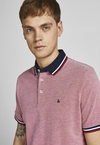 Rød tekstureret polo-shirt med en marineblå krave, der har hvide og røde striber, tre knapper og et lille logo på brystet.