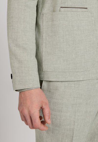 Zbliżenie na rękę obok kieszeni jasnoszarej lnianej marynarki i pasujących spodni.