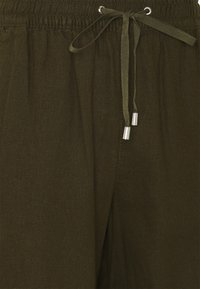 Zbliżenie na spodnie oliwkowe z zasuwanym pasem, z metalowymi końcówkami na końcach zawiązanych sznurków.