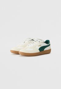 Weiße Sneaker mit grünen Akzenten, strukturiertem Stoff, dicker Gummisohle, runder Zehenpartie und auffälligem Logo auf der Zunge und der Seite.